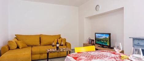 A living room with a sofa, a dining table set with plates and glasses, a television, and a clock on the wall.