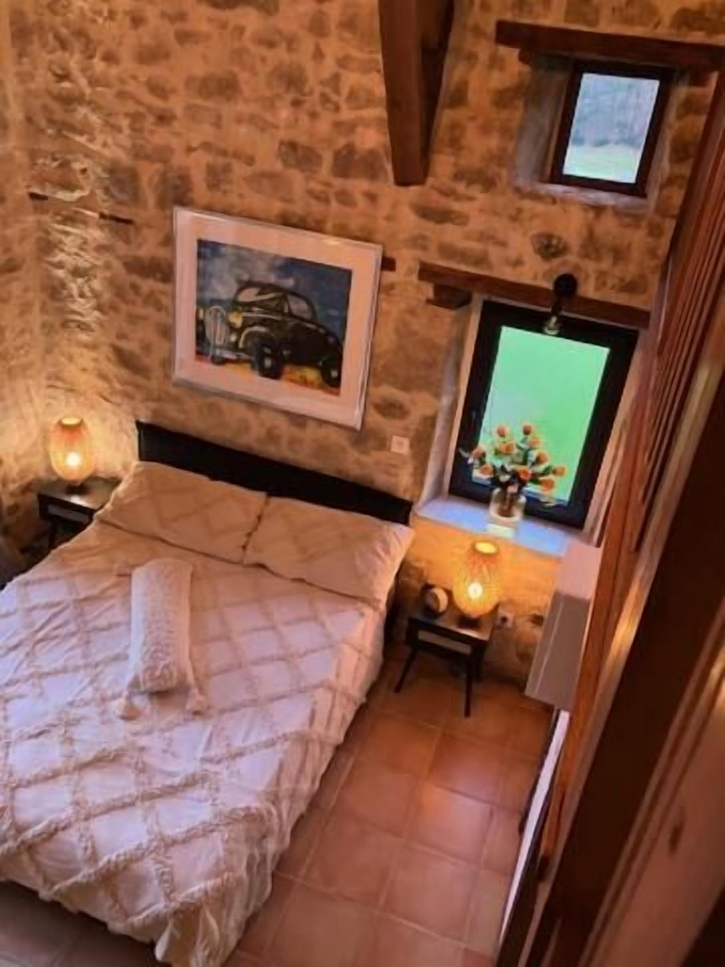 Un dormitorio con pared de piedra, cama con colcha, cuadro enmarco, ventana con vistas y mesita de noche con lámpara.