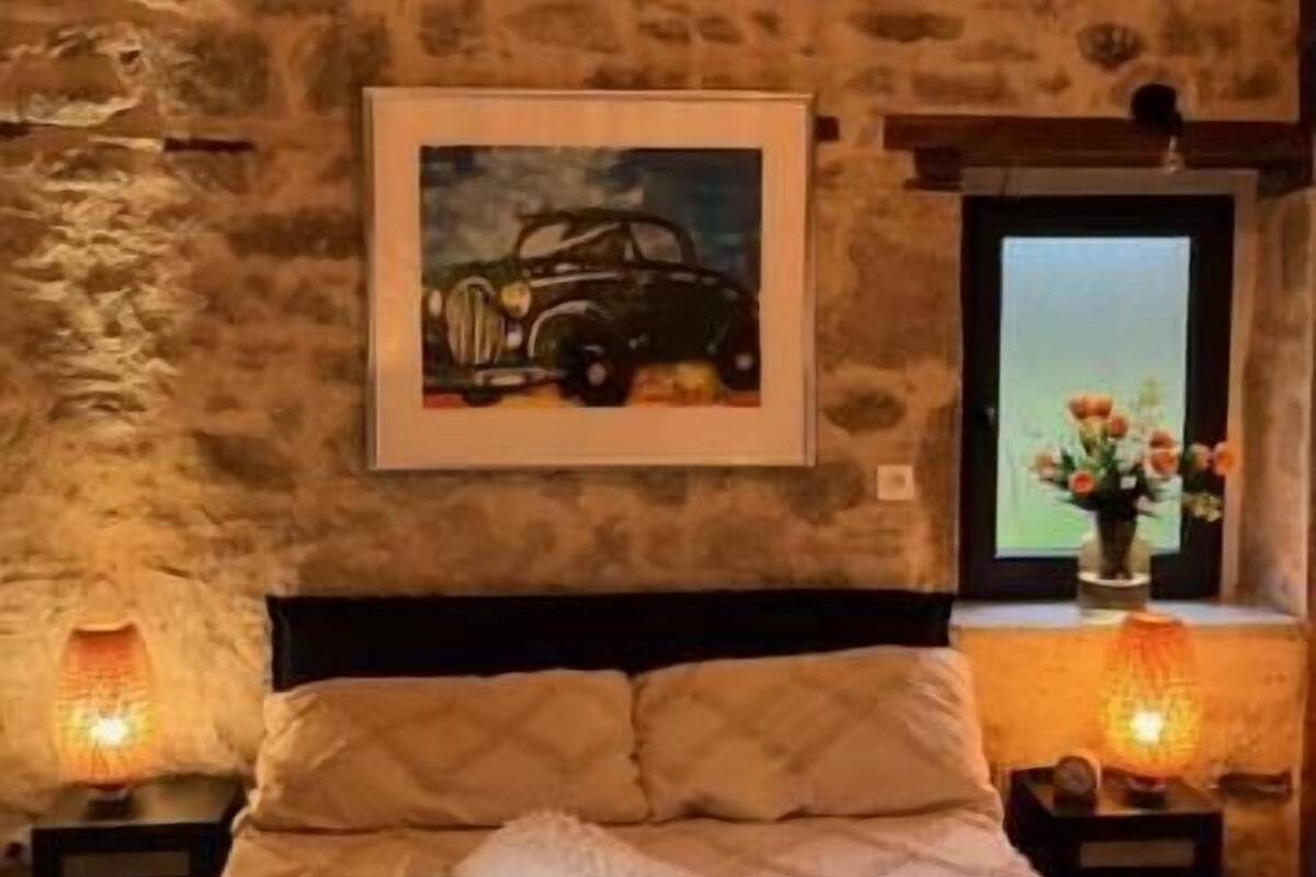 Une chambre avec un mur en pierre, un lit recouvert d’une courtepointe, un tableau représentant une voiture encadré, et deux lampes de chevet.