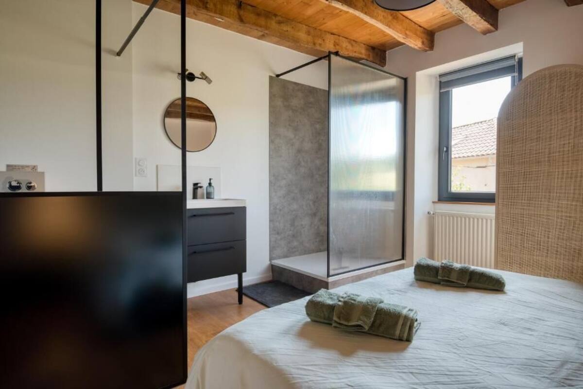 Une chambre moderne avec un lit, une douche à l’italienne, un miroir rond et une fenêtre.