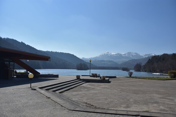 A lakeside view with a mountain backdrop, a concrete walkway, and a building with a sloped roof. - Domaine du Lac Chambon (Murol)