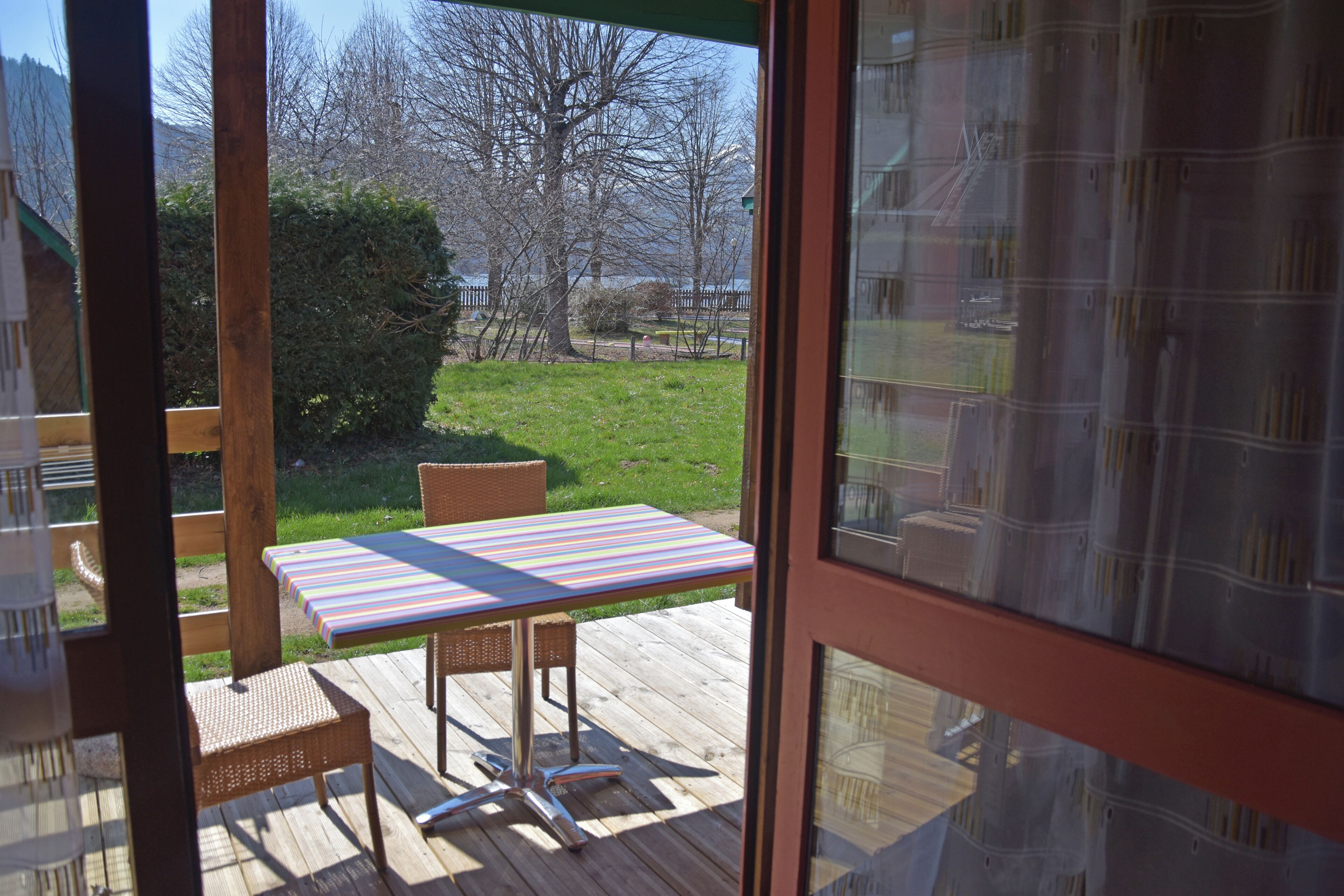 A wooden deck with a table and chairs, a view of a grassy area with trees, and a body of water in the background.