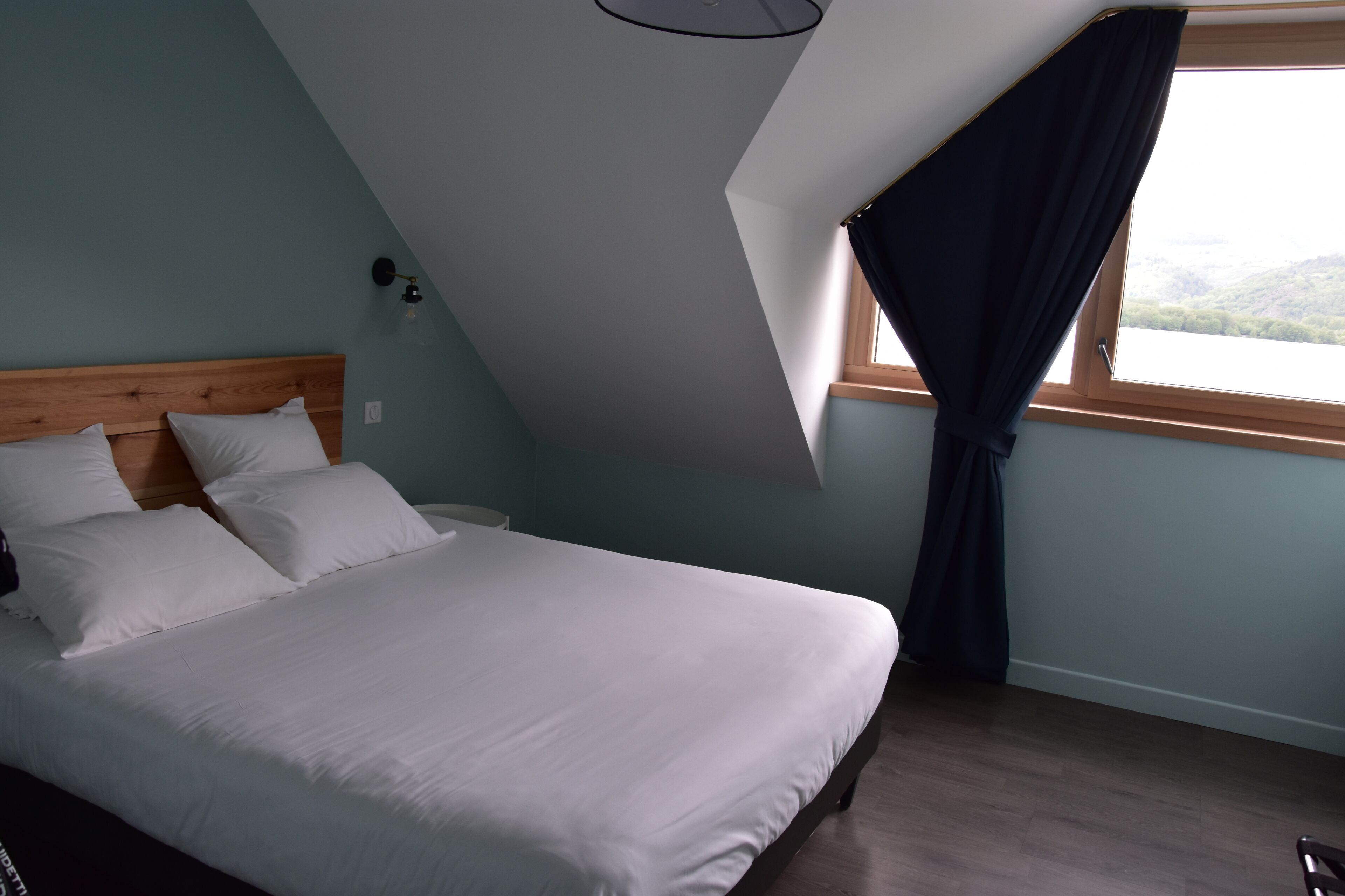 Une chambre avec un plafond en pente, un lit recouvert de draps blancs, une table de chevet, une lampe fixée au mur et une fenêtre avec un rideau.