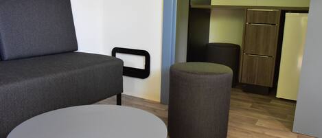 Ein gemütlicher, kleiner Wohnraum mit grauem Sofa, rundem Tisch, Rattan-Pouff, einer Küchenzeile und einem Fenster mit Blick auf Grünflächen.