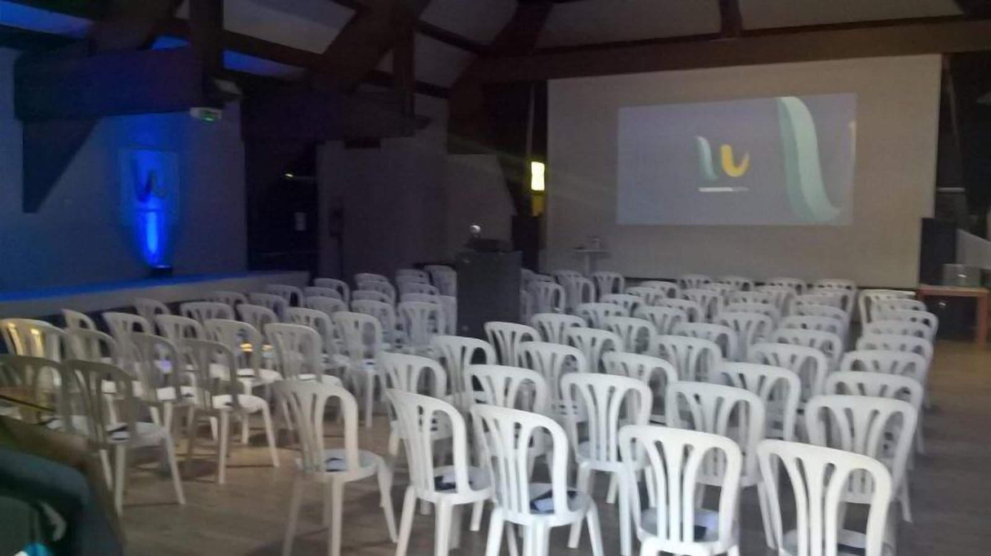 Un salón de conferencias con hileras de sillas de plástico blanco, una pantalla grande y un escenario con iluminación azul.