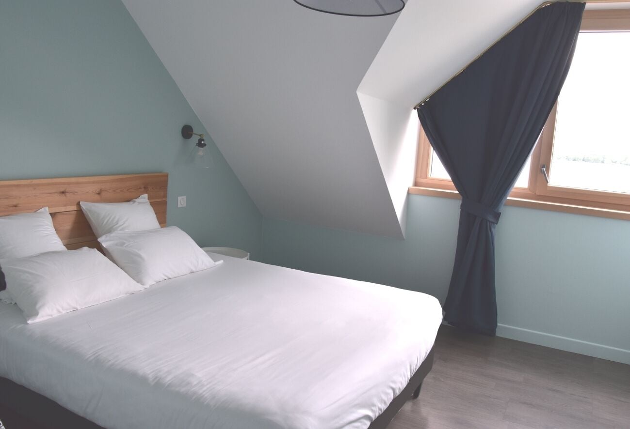 A bedroom with a sloped ceiling, a bed with white bedding, a window with curtains, and a wall-mounted light fixture.