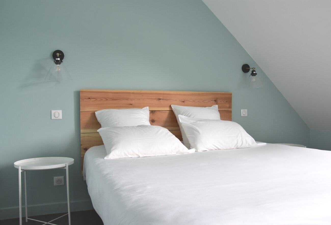 A bedroom with a wooden headboard, white bedding, a small white side table, and two wall-mounted light fixtures.