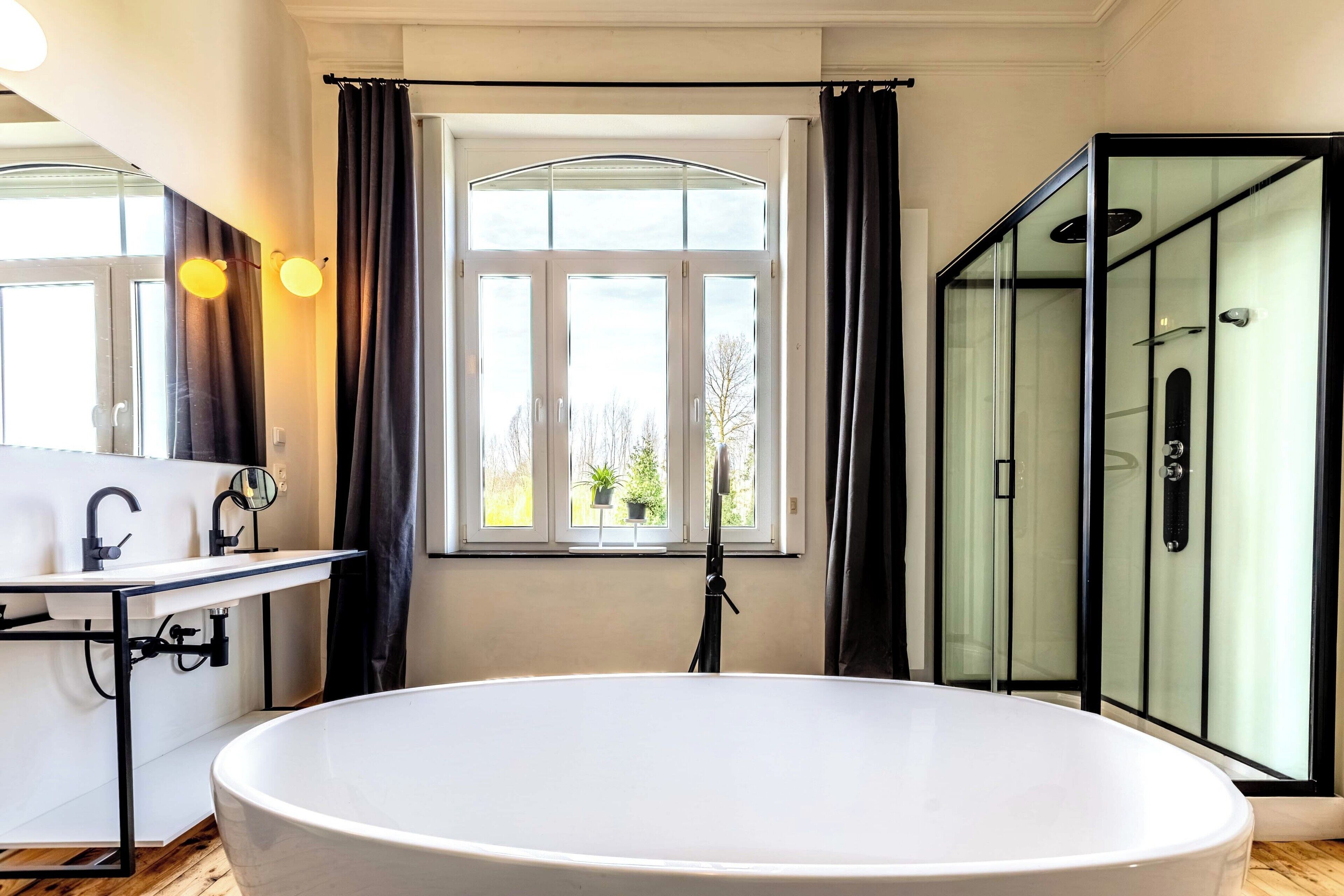 A bathroom with a freestanding bathtub, a window with curtains, a glass shower enclosure, and a sink with a mirror.