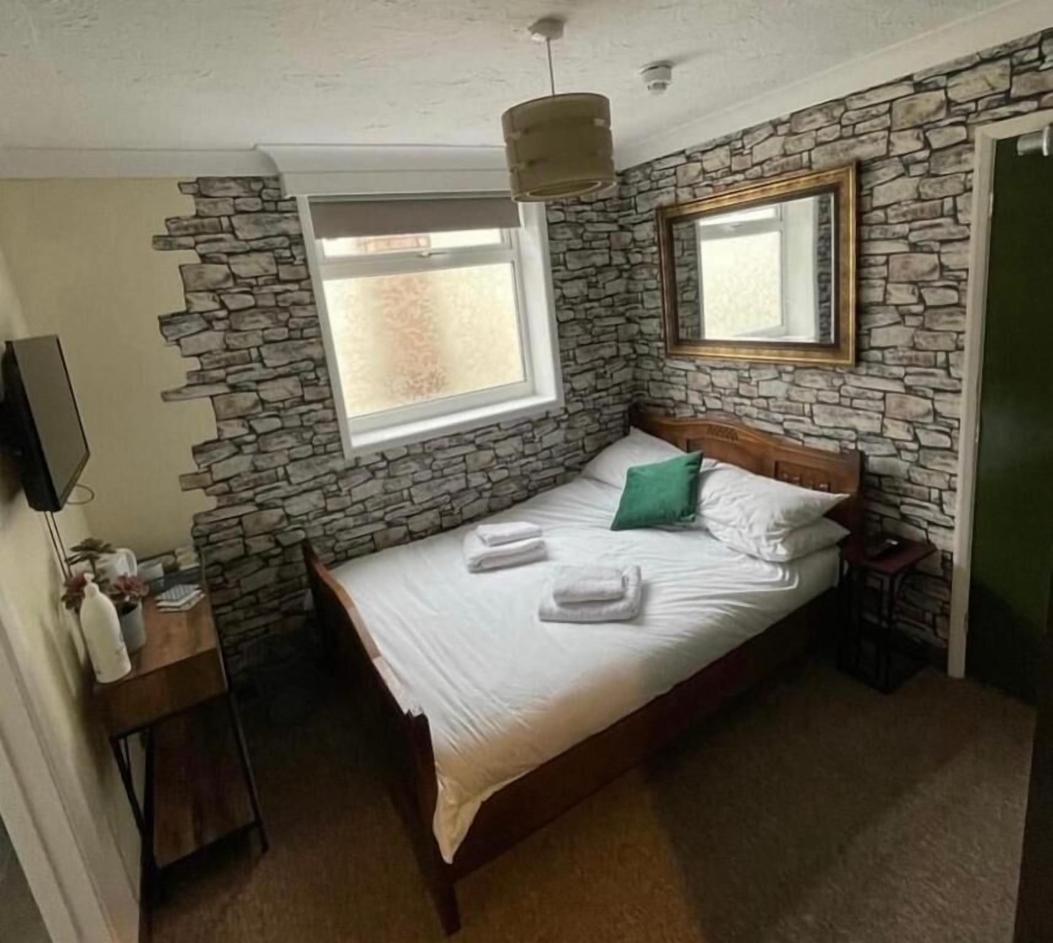 A bedroom with a stone wall, a bed with white linens, a green pillow, a bedside table, and a mirror.