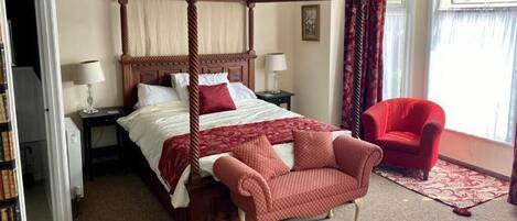 A four-poster bed with a red and white bedspread, a red armchair, a nightstand with a lamp, and a window with curtains.
