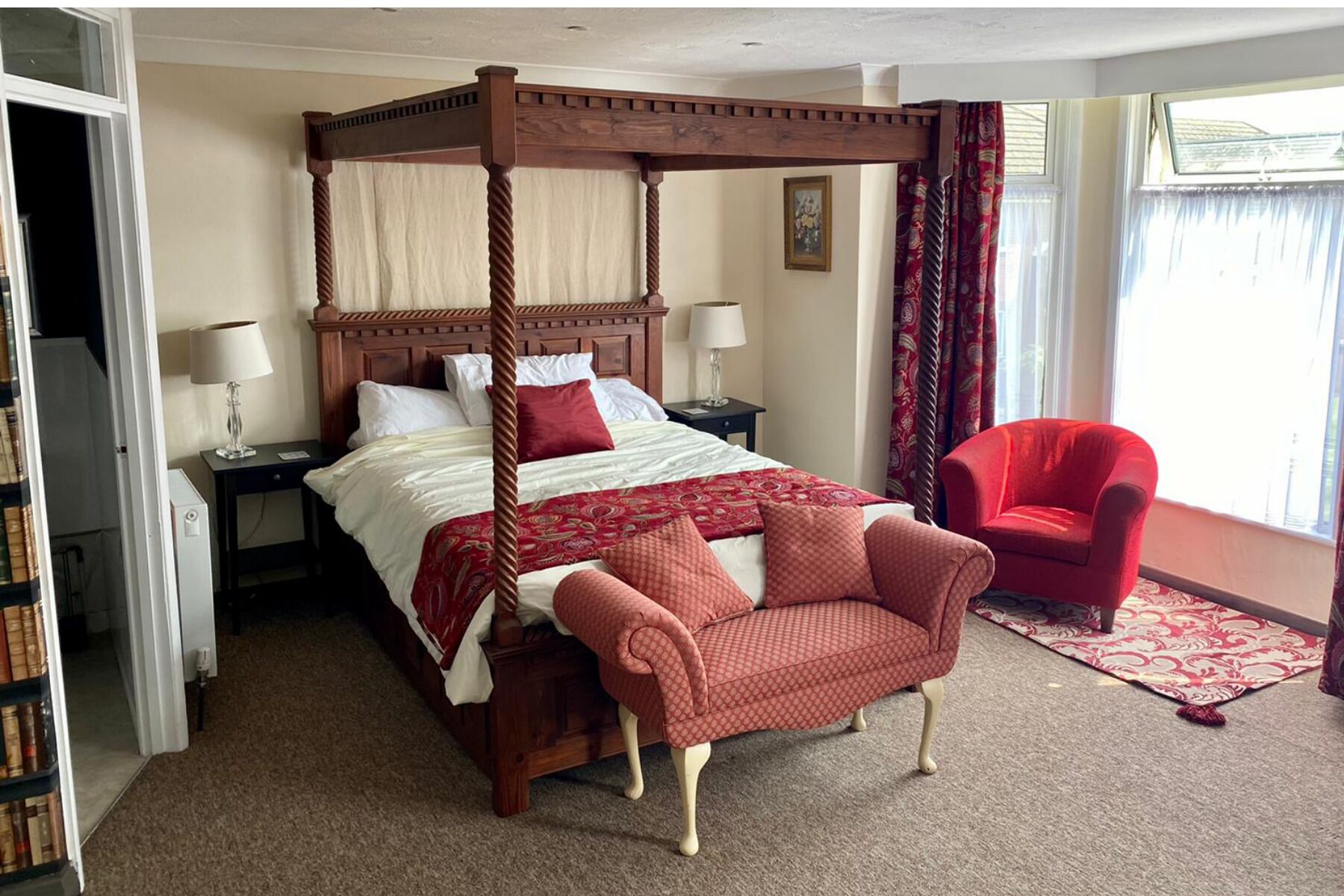 A four-poster bed with a red and white bedspread, a red armchair, a nightstand with a lamp, and a window with curtains.