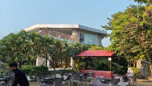 An outdoor seating area with tables and chairs, a building in the background, and a person serving food. - The Manrals Resort (Ramnagar)