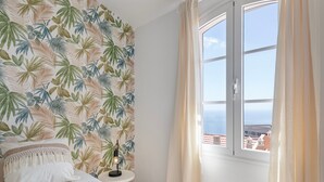 A bedroom with a bed, a nightstand, a lamp, a window with curtains, and a tropical wallpaper.