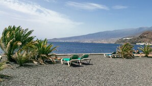 Ein Kiesstrand mit Liegestühlen und Palmen, mit Blick auf eine Küstenstadt und Berge.