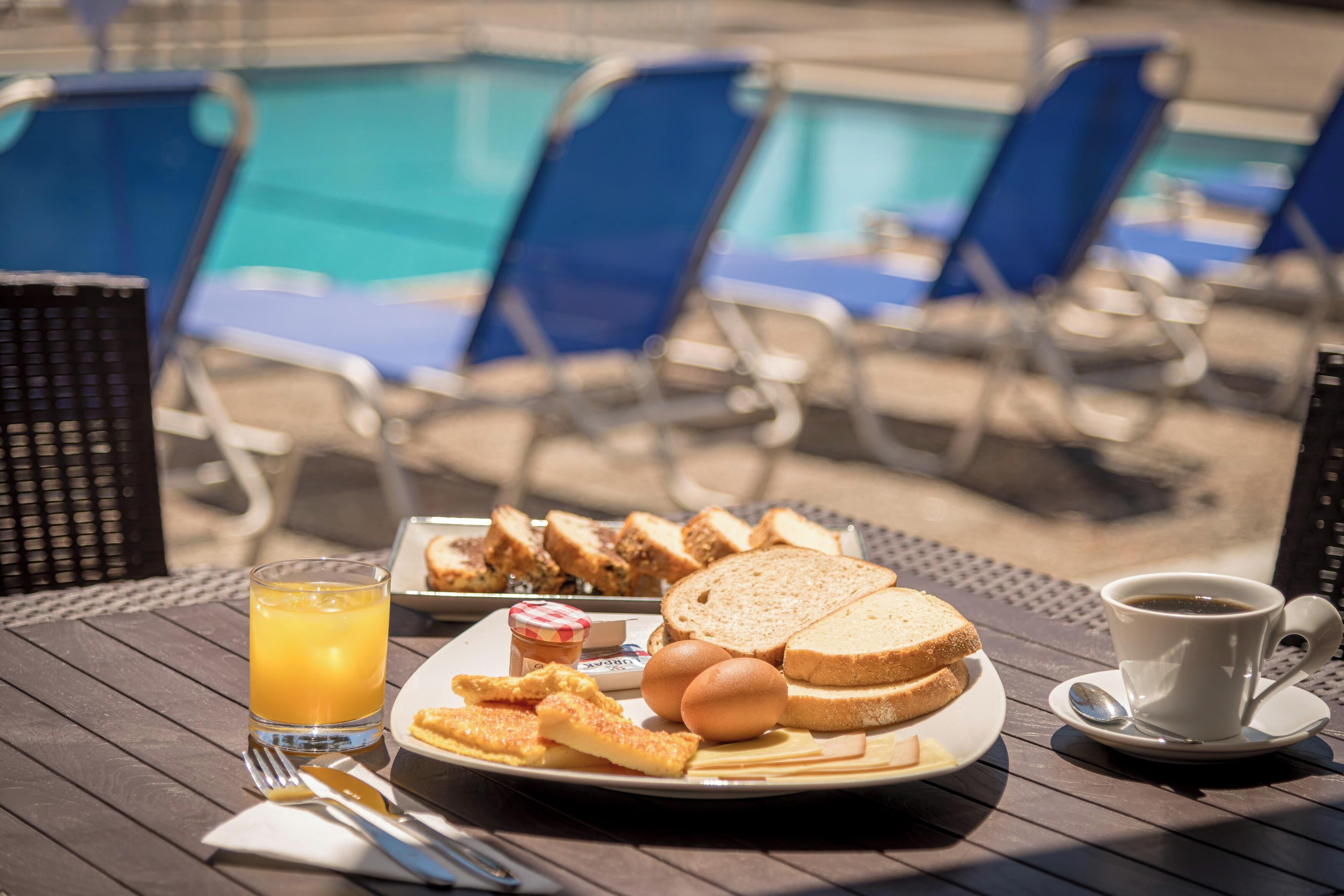 A breakfast tray with toast, eggs, and a glass of orange juice on a wooden table by a poolside.