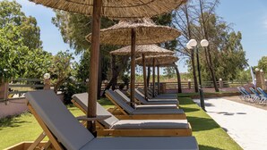 A row of wooden lounge chairs with thatched umbrellas, arranged on a grassy area with trees in the background. - Apollo Hotel (Zakynthos)