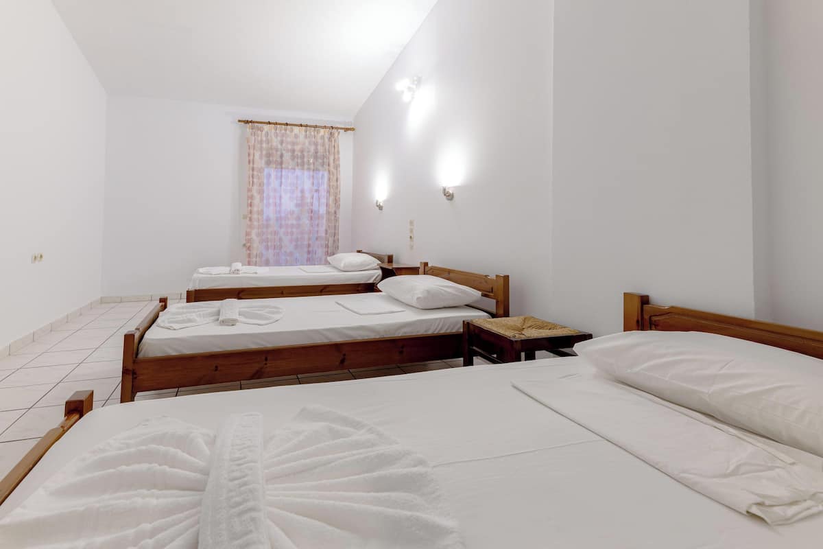 A room with three wooden beds, white linens, a window with a curtain, and two wall-mounted lights.