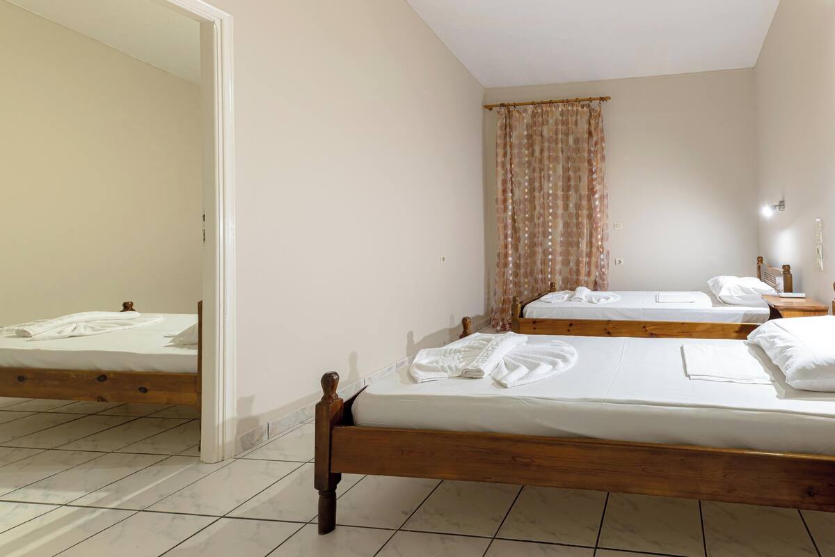 A bedroom with two single beds, a wooden headboard, a mirror, a window with curtains, and a tiled floor.