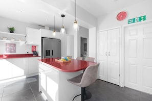 A modern kitchen with white cabinets, a red countertop, pendant lights, a clock, and a fire exit sign. - Spacious 5 Bedroom Family Home in Harrow (Harrow)