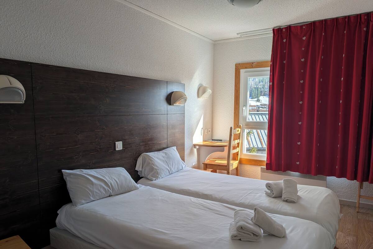 Ein Hotelzimmer mit zwei Betten, einem Holzkopfstück, einem Schreibtisch, einem Stuhl, einem Fenster mit roten Vorhängen und Blick auf ein Gebäude.