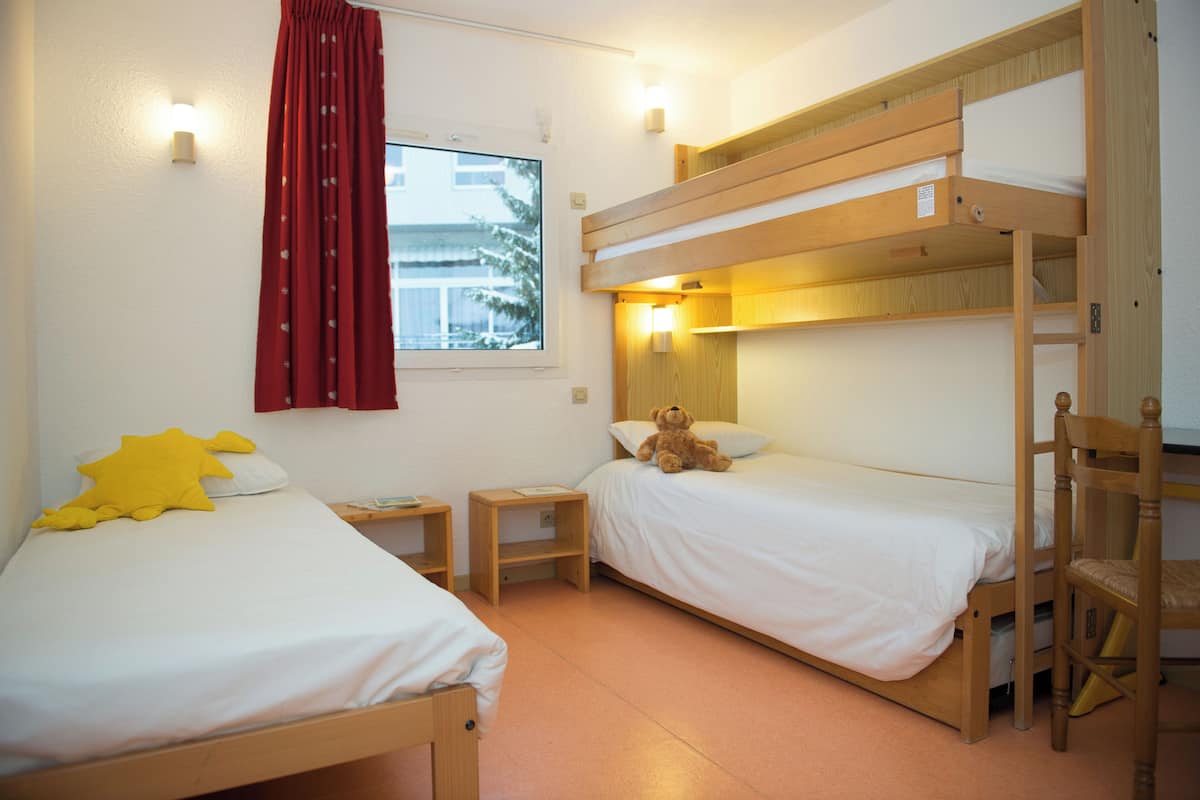Ein Zimmer mit zwei Etagenbetten, einem Holzbstuhl, einem kleinen Nachttisch und einem Fenster mit rotem Vorhang.