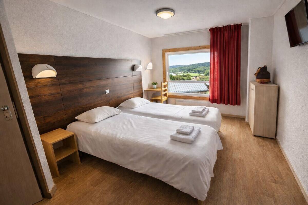 Ein Hotelzimmer mit zwei Betten, einem hölzernen Kopfteil, einem kleinen Tisch, einem Nachttisch, einem Fernseher und einem Fenster mit rotem Vorhang.