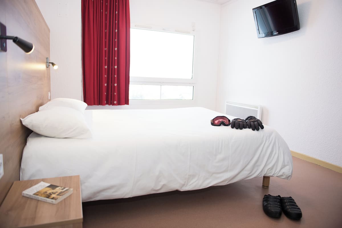 Ein Hotelzimmer mit einem Bett, einem an der Wand montierten Fernseher, einem Fenster mit roten Vorhängen und einem Paar Schuhe auf dem Boden.
