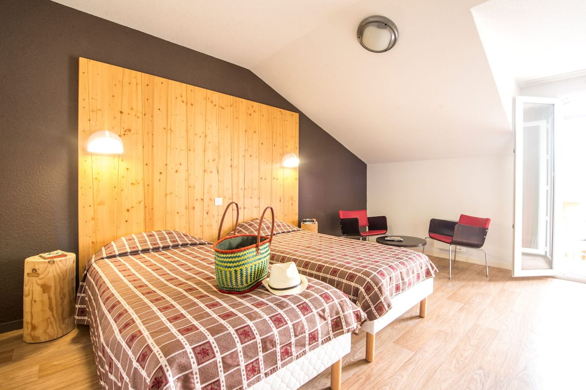 Ein Zimmer mit zwei Betten, einem hölzernen Kopfteil, einem kleinen Tisch, einem Stuhl und einer Tür, die auf einen Balkon führt.