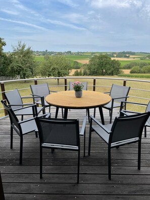Ein runder Holztisch mit sechs Stühlen auf einer Terrasse, mit Blick auf eine ländliche Gegend.