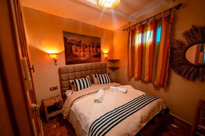 A hotel room with a bed, a painting on the wall, a wall-mounted lamp, a striped pillow, a towel, and a mirror. - Riad Bleu Azahraa (Safi)