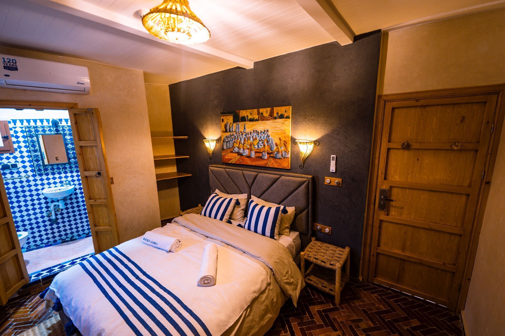 Habitación de hotel con cama, aire acondicionado de pared, un cuadro, puerta de madera y suelo de baldosas.