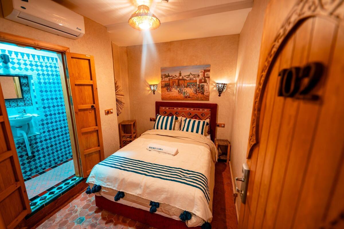 A hotel room with a bed, a bathroom with a sink, a door with a number, and a wall-mounted lamp.