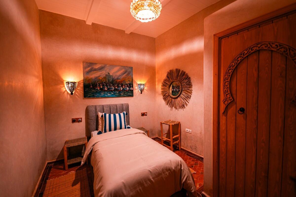A bedroom with a bed, a painting, wall sconces, a decorative mirror, and a wooden door.