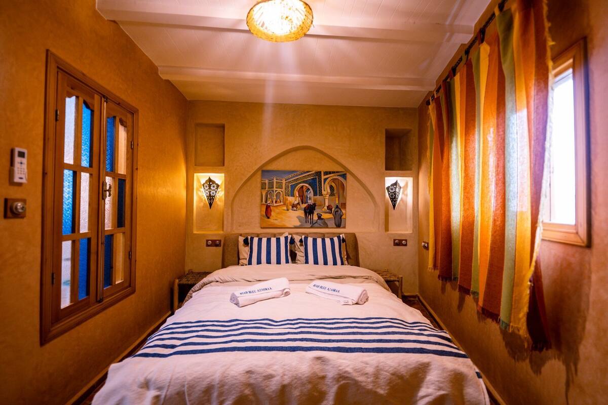 A hotel room with a bed, a painting above the bed, a door with a window, and a wall-mounted light fixture.