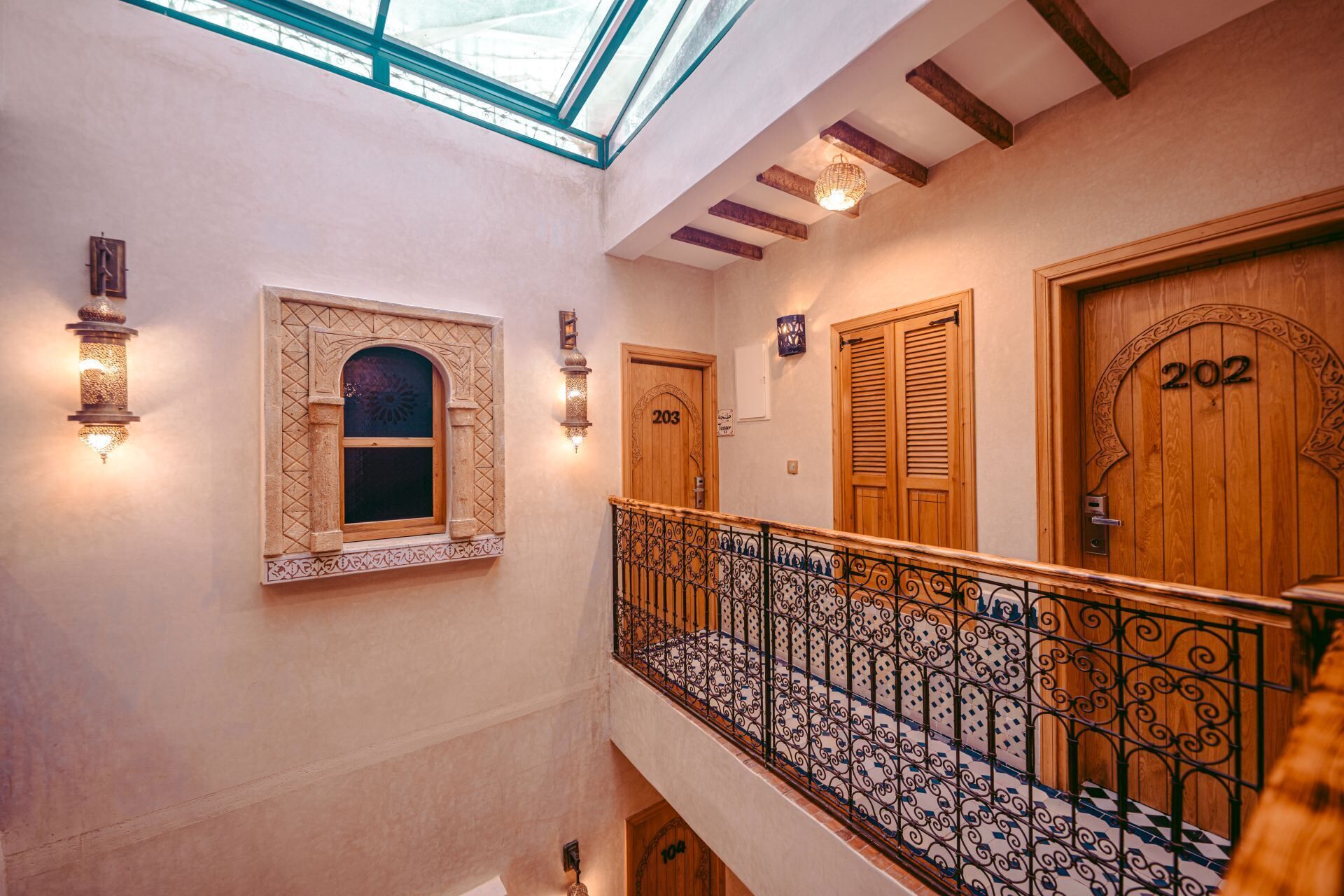 A hotel interior with a skylight, wooden doors numbered 203 and 202, and a decorative wrought iron railing.