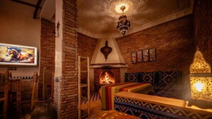 A cozy room with a brick fireplace, a sofa with patterned cushions, a mounted painting, and a hanging light fixture. - Riad Bleu Azahraa (Safi)