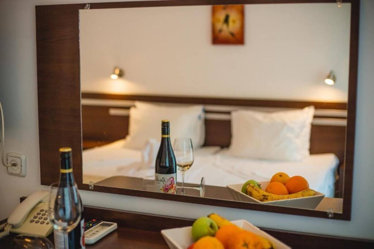 A hotel room with a bed, a bedside table with a phone, a bottle, a glass, and a tray with fruit.