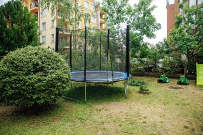 Un trampolín en un área con césped, bordeado por un seto, árboles y un edificio al fondo.