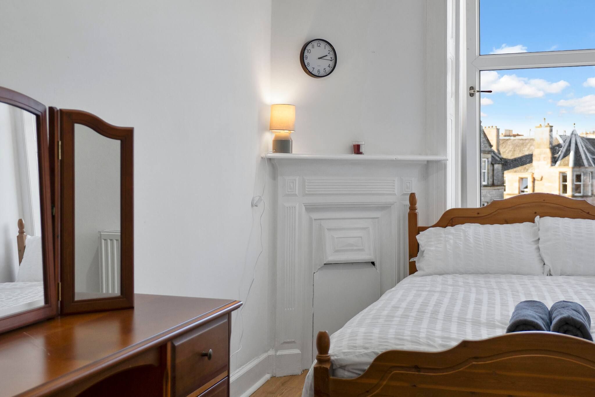 A bedroom with a wooden bed, a white dresser, a mirror, a wall clock, and a window with a city view.