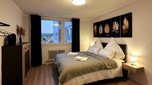Een slaapkamer met een bed, een nachtkastje met een lamp, een radiator, een raam met gordijnen, en een wand met bladkunst.