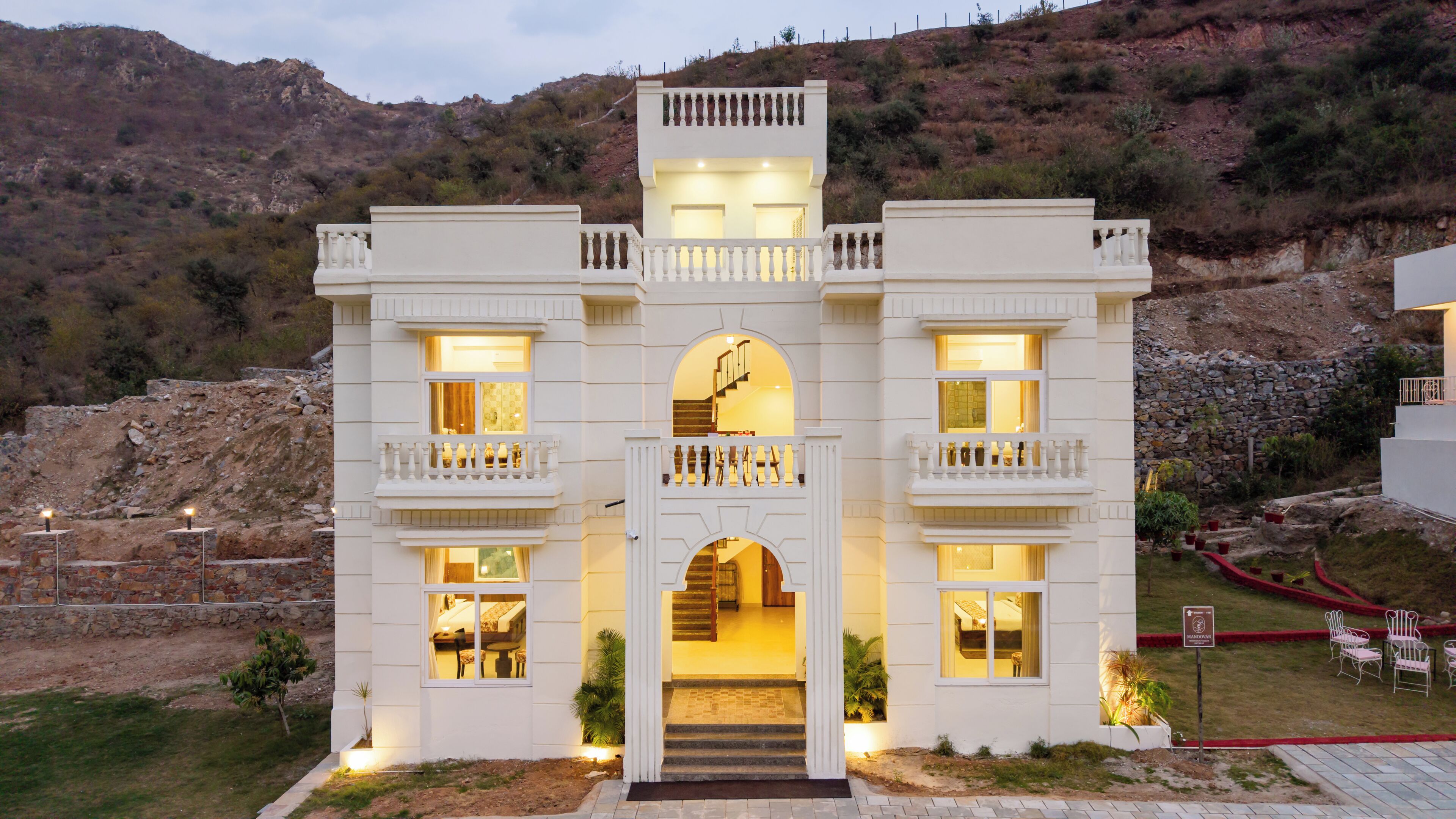 A white building with multiple balconies, a central staircase, and a stone wall in the background.
