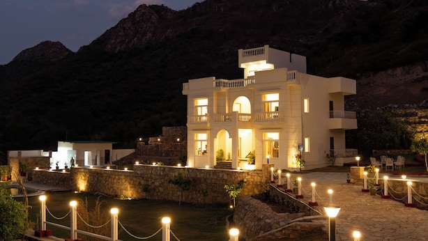 A white building with multiple balconies and arched windows, surrounded by a stone wall and a pool area. - ELIVAAS Mandovar Mountain Valley Retreat (Udaipur)