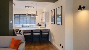 Une cuisine moderne avec des luminaires suspendus, un coin repas doté de tabourets de bar et un canapé agrémenté d’un coussin.