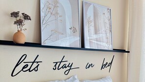 A bed with decorative pillows, a wall-mounted shelf with framed artwork, and a wall decal with the text 'lets stay in bed'. - Pearly EuroPoint (Saint-Louis)