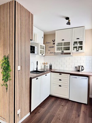 A modern kitchen with white cabinetry, a wooden accent wall, a microwave, a sink, and a coffee maker. - Pearly EuroPoint (Saint-Louis)