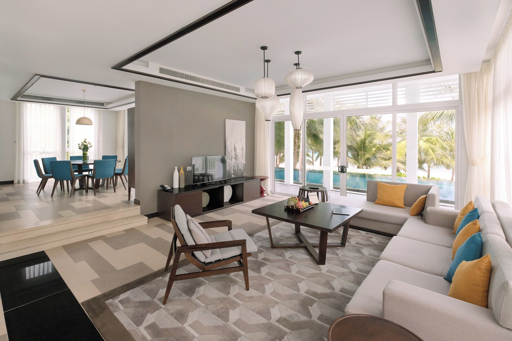 Una sala moderna con sofá, mesa de centro y sillas. Hay un comedor con mesa y sillas. La habitación tiene ventanas grandes con vista a palmeras y una piscina.
