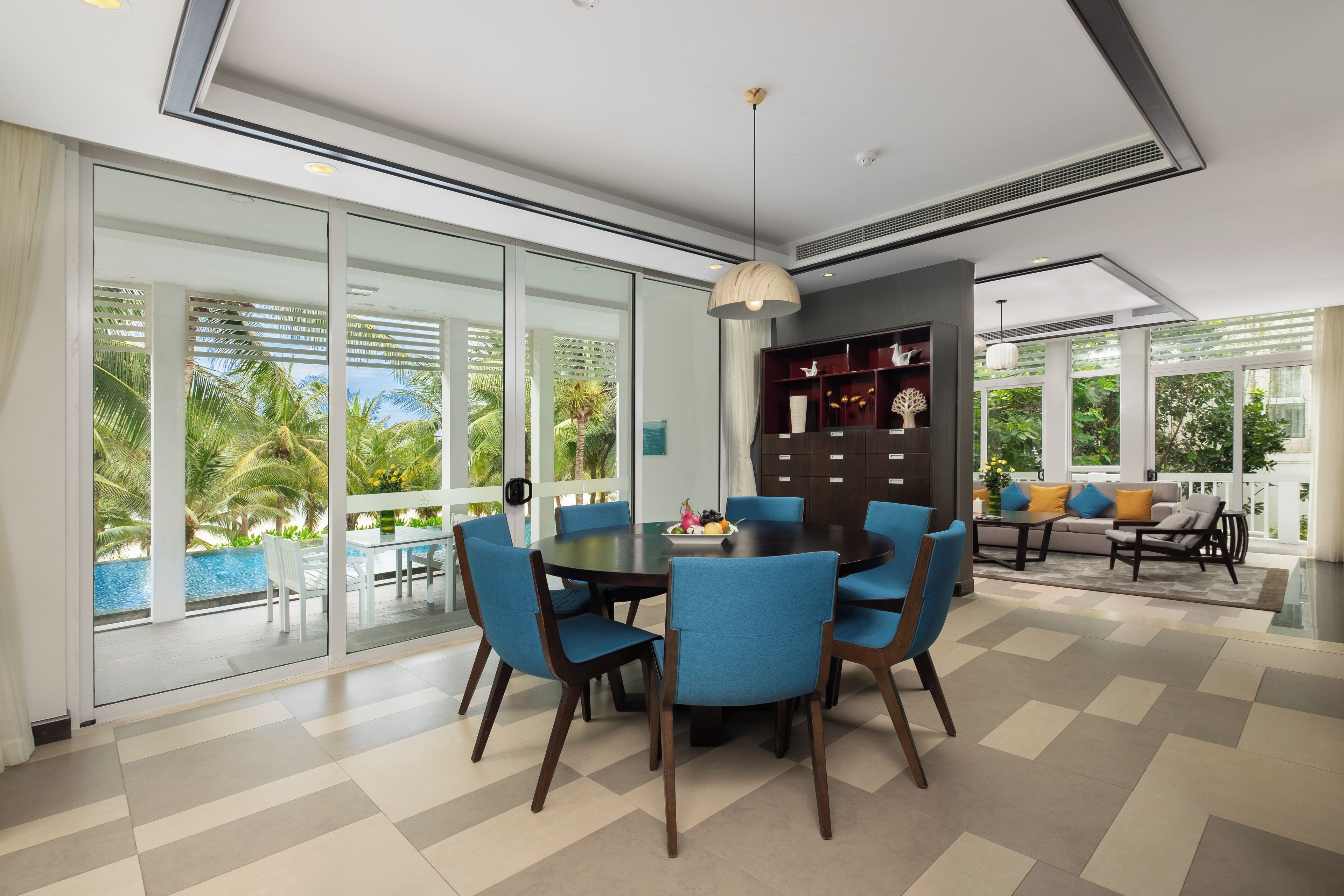 A modern dining area with a round table, blue chairs, and a view of a pool and palm trees through large windows.