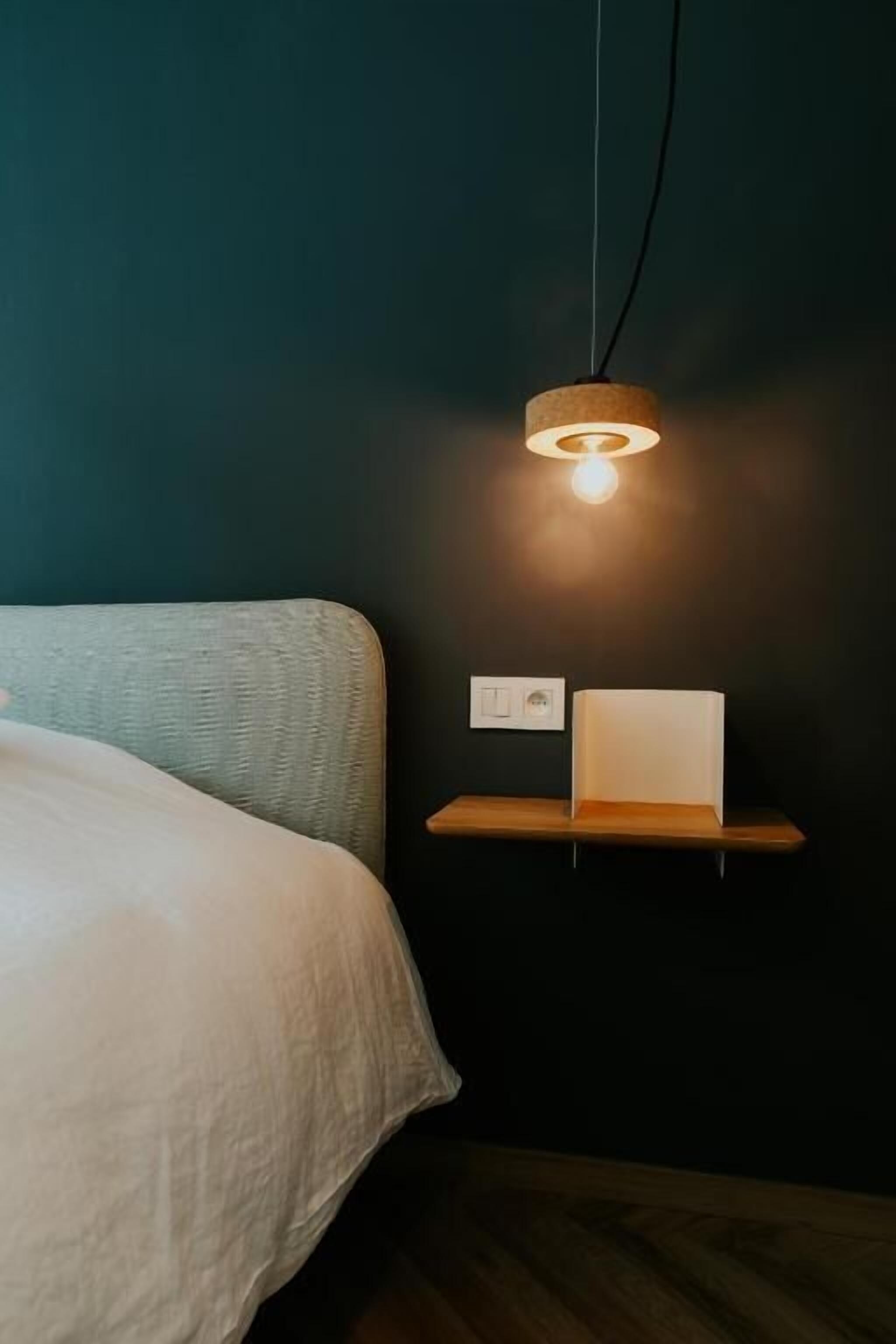 Een bed met een witte deken, een hanglamp en een klein wit schapje tegen een donkere muur.