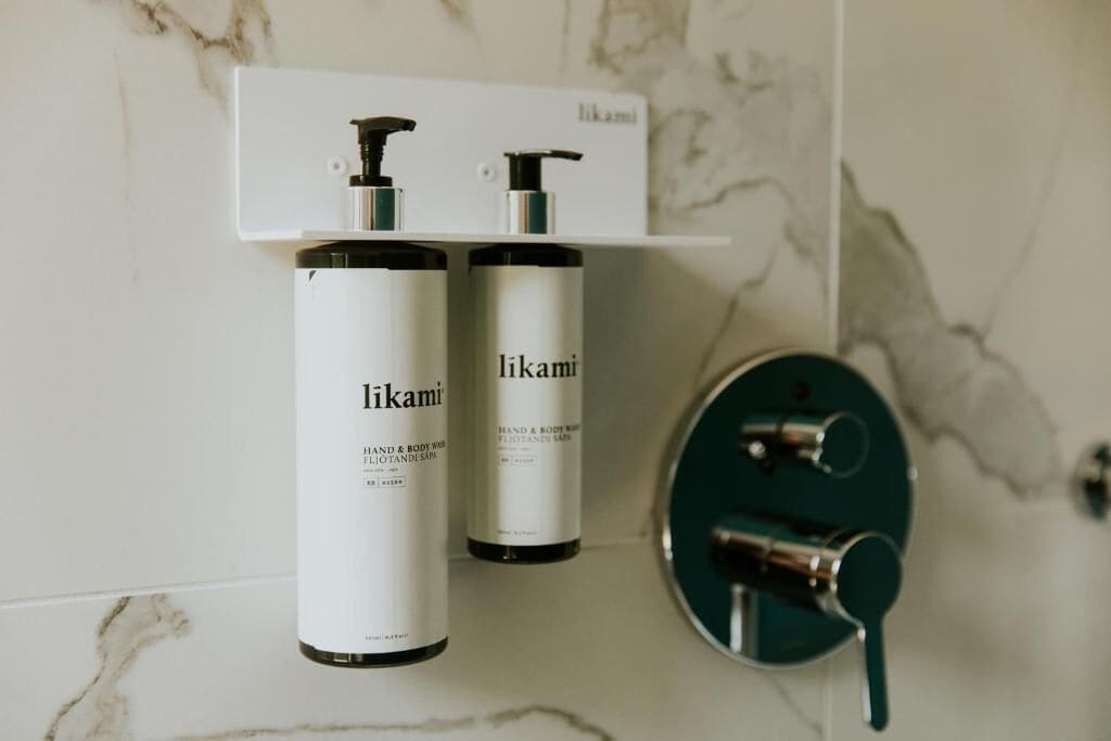Two bottles of Likami hand and body wash on a white shelf in a bathroom with a marble wall.