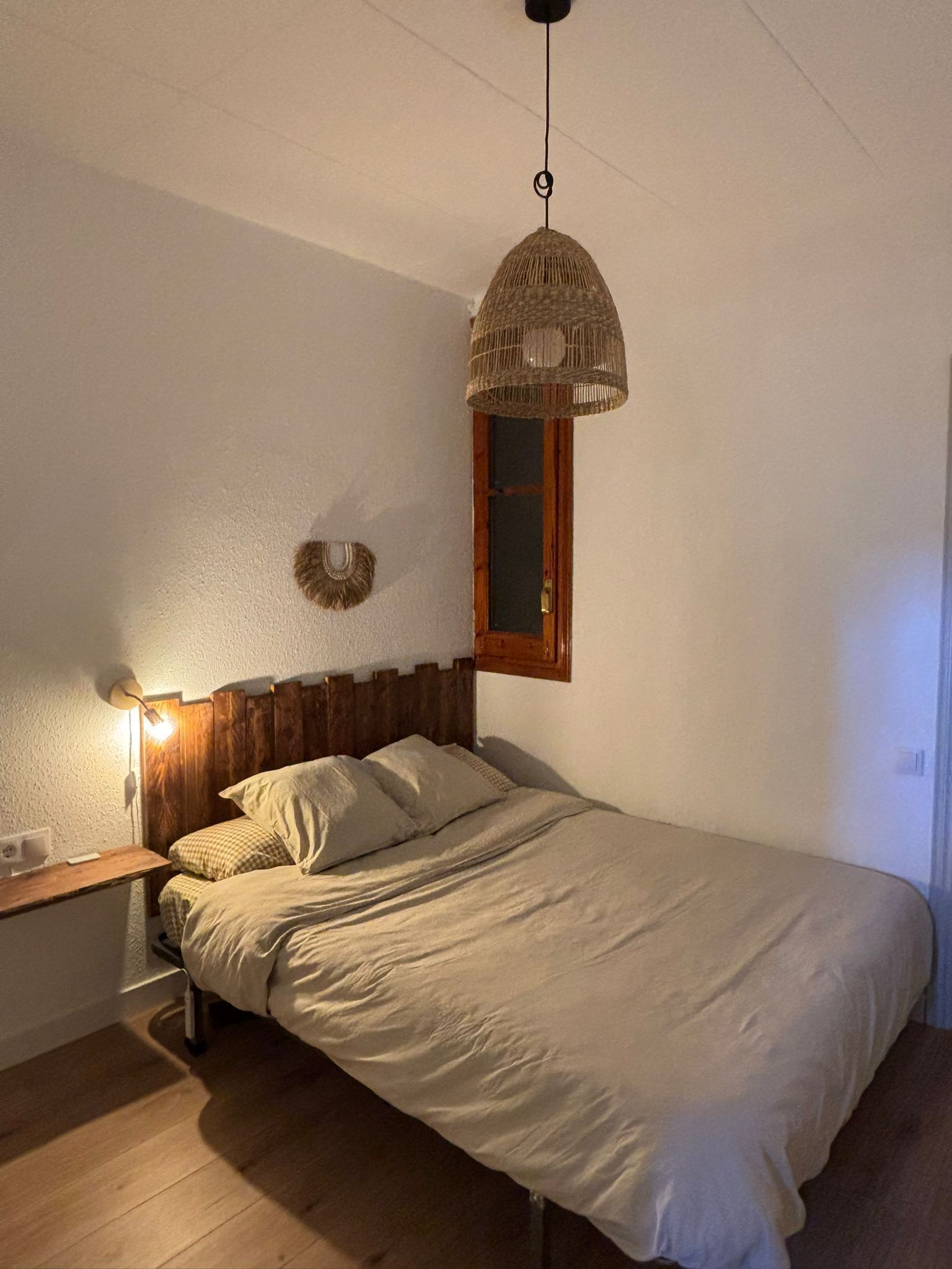 A bedroom with a wooden headboard, a single bed, a bedside table, a wall-mounted lamp, and a hanging wicker lamp.